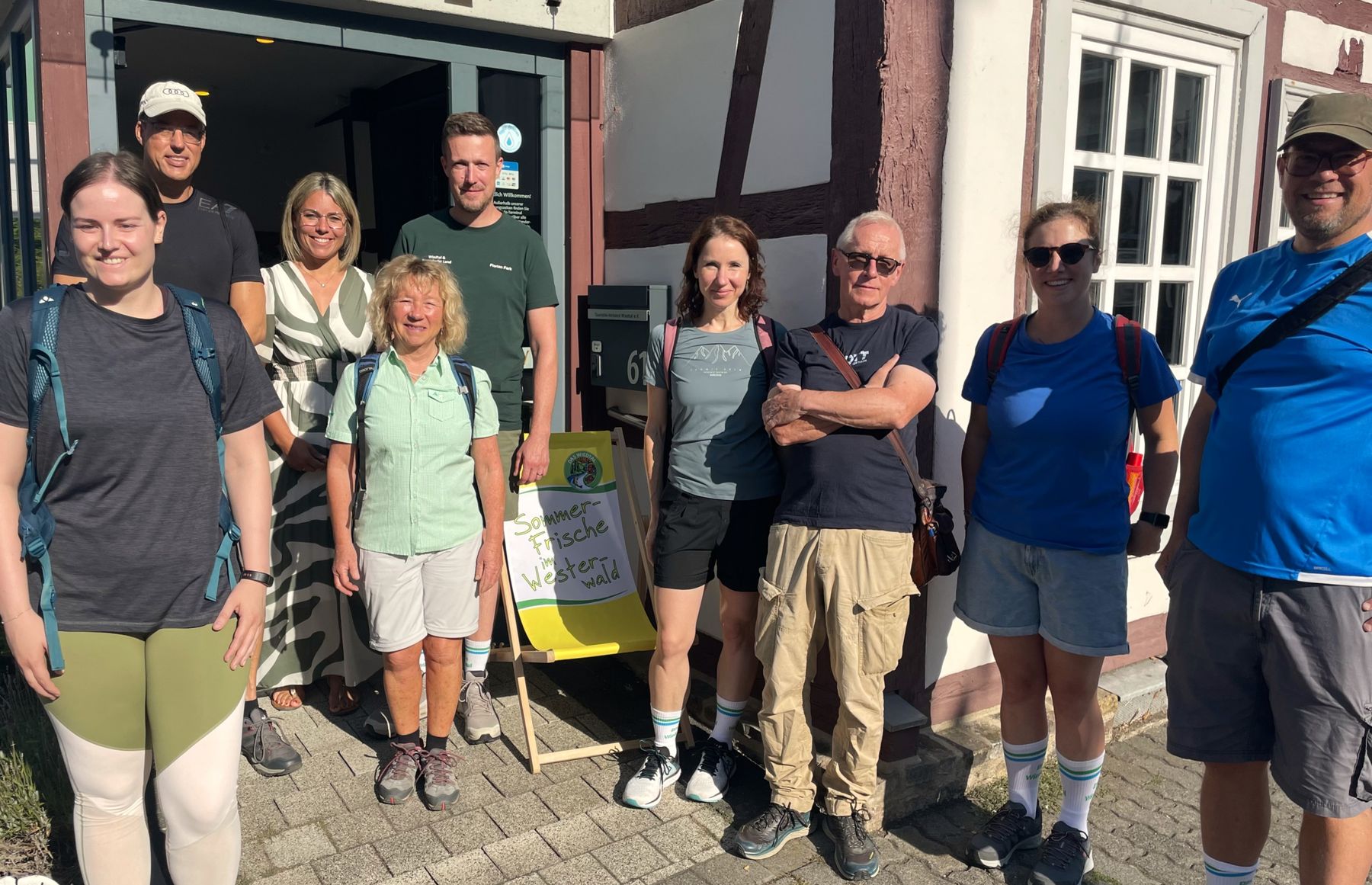 Nine people stand smiling in front of a building on a sunny day, some wearing backpacks and outdoor clothing, with a sign reading Westliche Wälder visible behind them near the entrance.